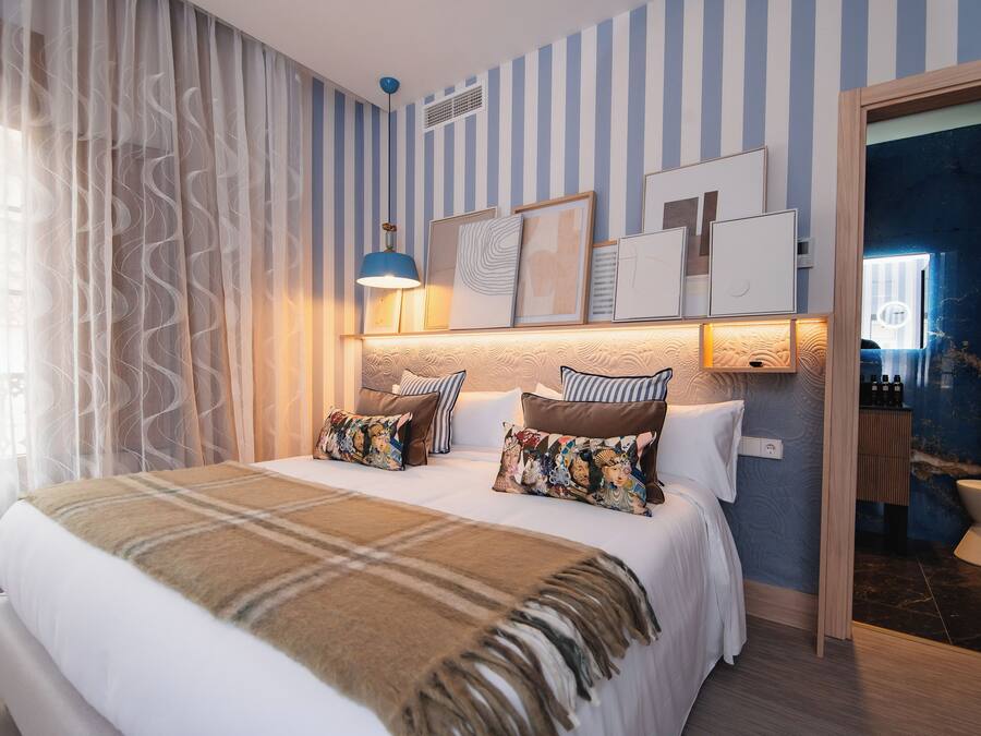 A hotel room with a bed, a bedside lamp, a window with curtains, a wall with striped wallpaper, and a door leading to a bathroom.