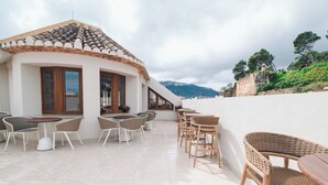 A terrace with wicker chairs and tables, a white building with a tiled roof, and a view of a hill with trees.