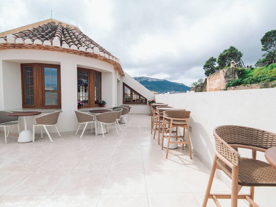 A terrace with wicker chairs and tables, a white building with a tiled roof, and a view of a hill with trees.