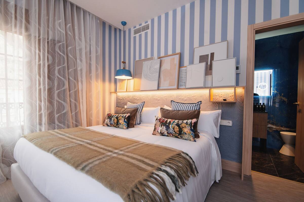 A hotel room with a bed, a bedside lamp, a window with curtains, a wall with striped wallpaper, and a door leading to a bathroom.