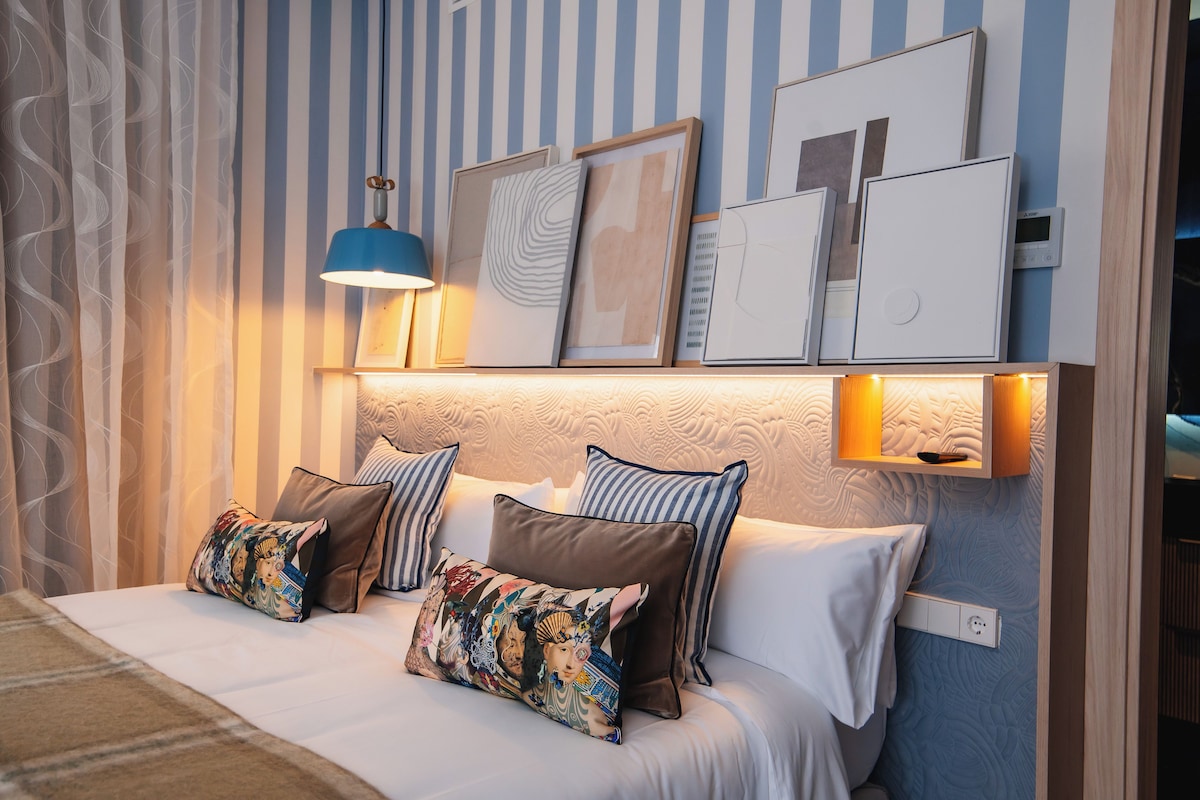 Ein ordentlich gemachtes Bett mit vielen Kissen, einer Wandlampe und gerahmten Bildern.