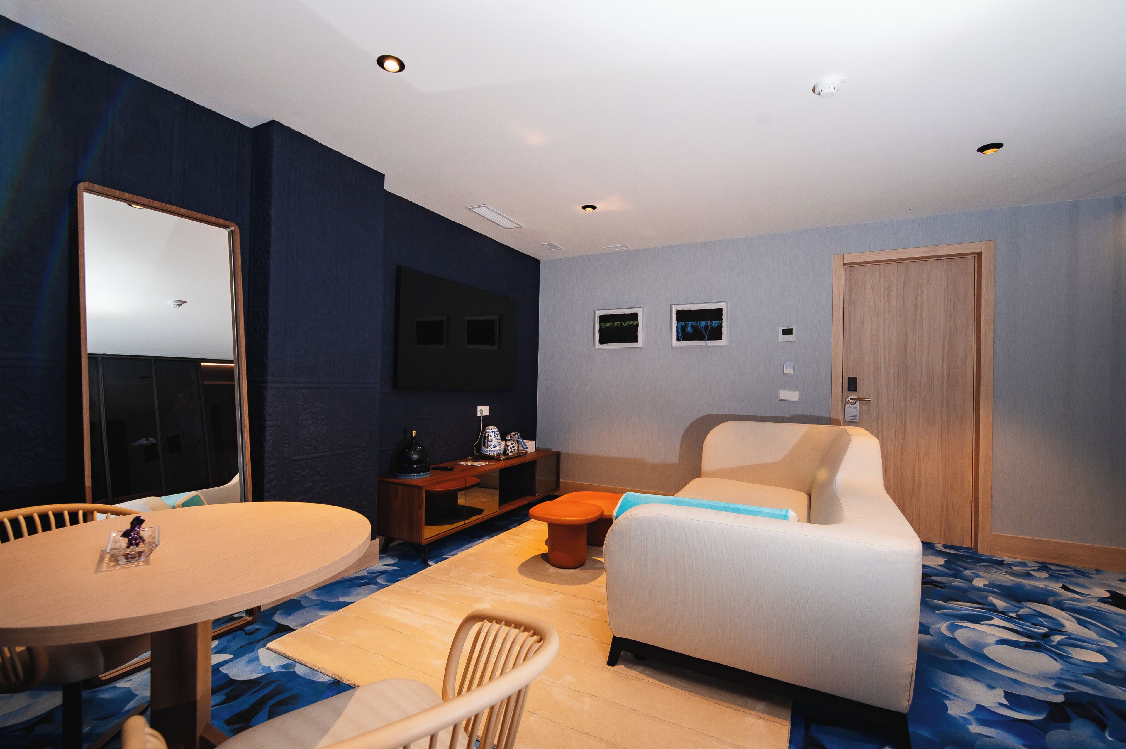 A hotel room with a round dining table, a sofa, a TV, and a wooden door.