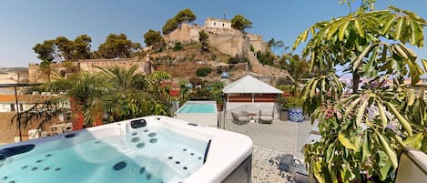 Una terraza en la azotea con jacuzzi, mobiliario exterior y vistas a una estructura histórica en la cima de una colina.
