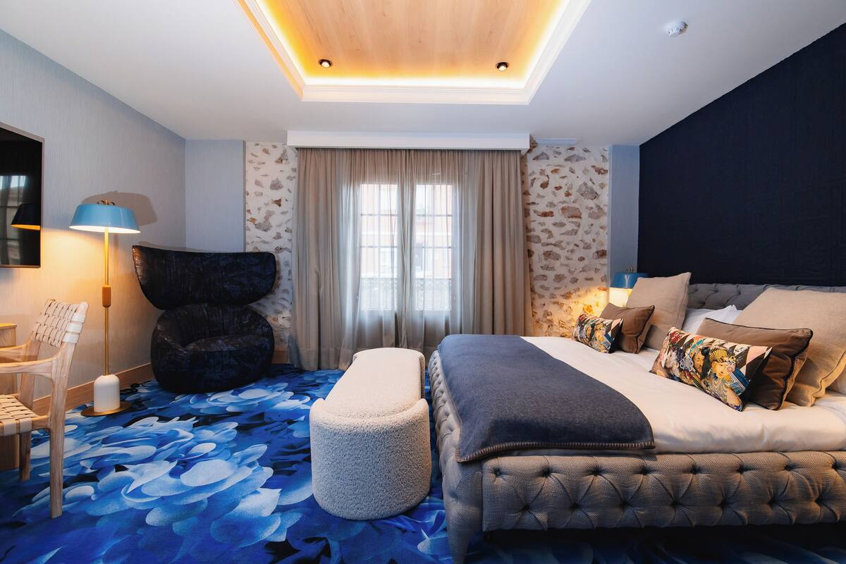 A modern hotel room with a large bed, a chair, a small ottoman, and a desk with a lamp.