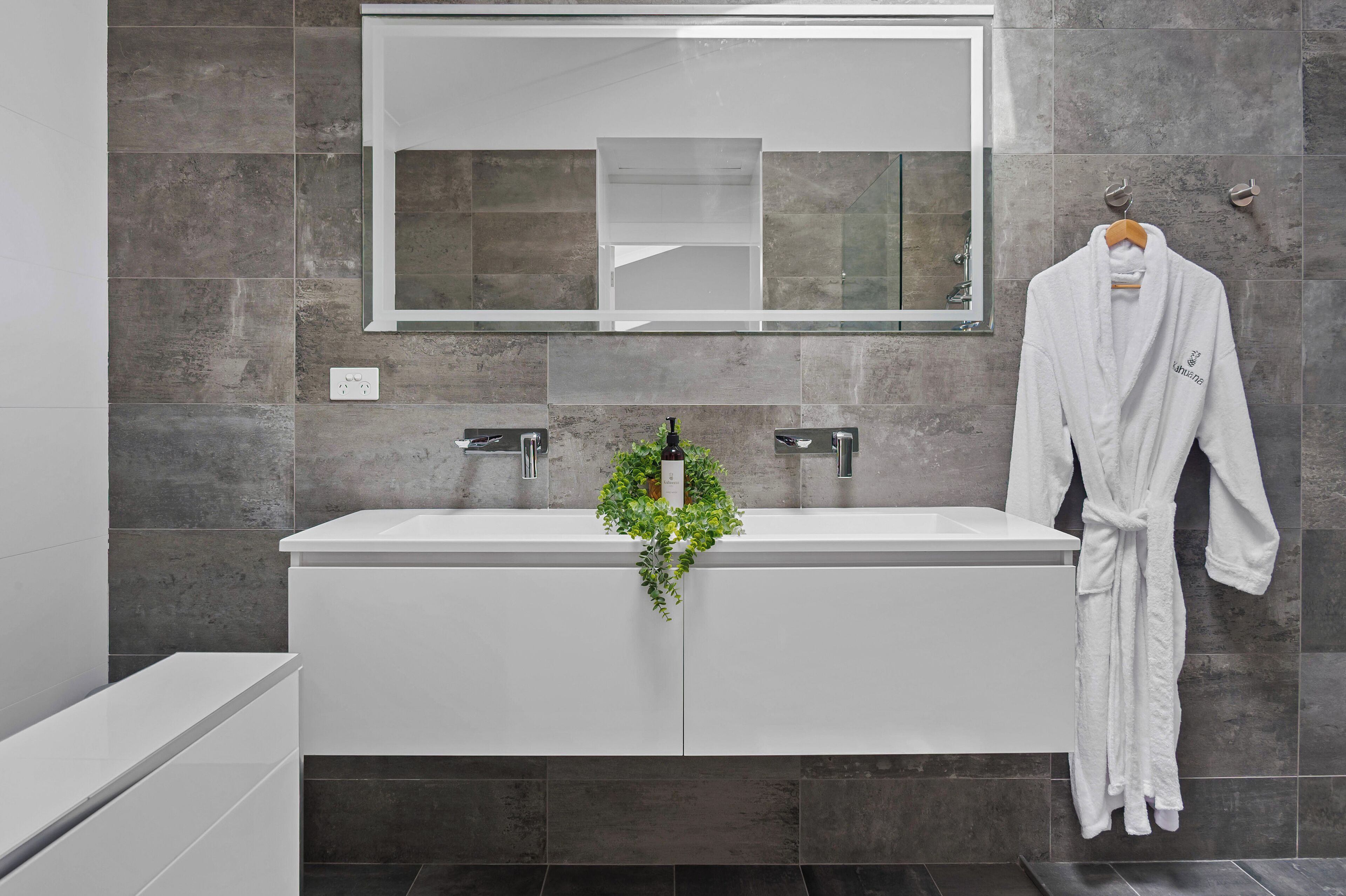 A modern bathroom with a large mirror, two sinks, and a robe hanging on the wall.