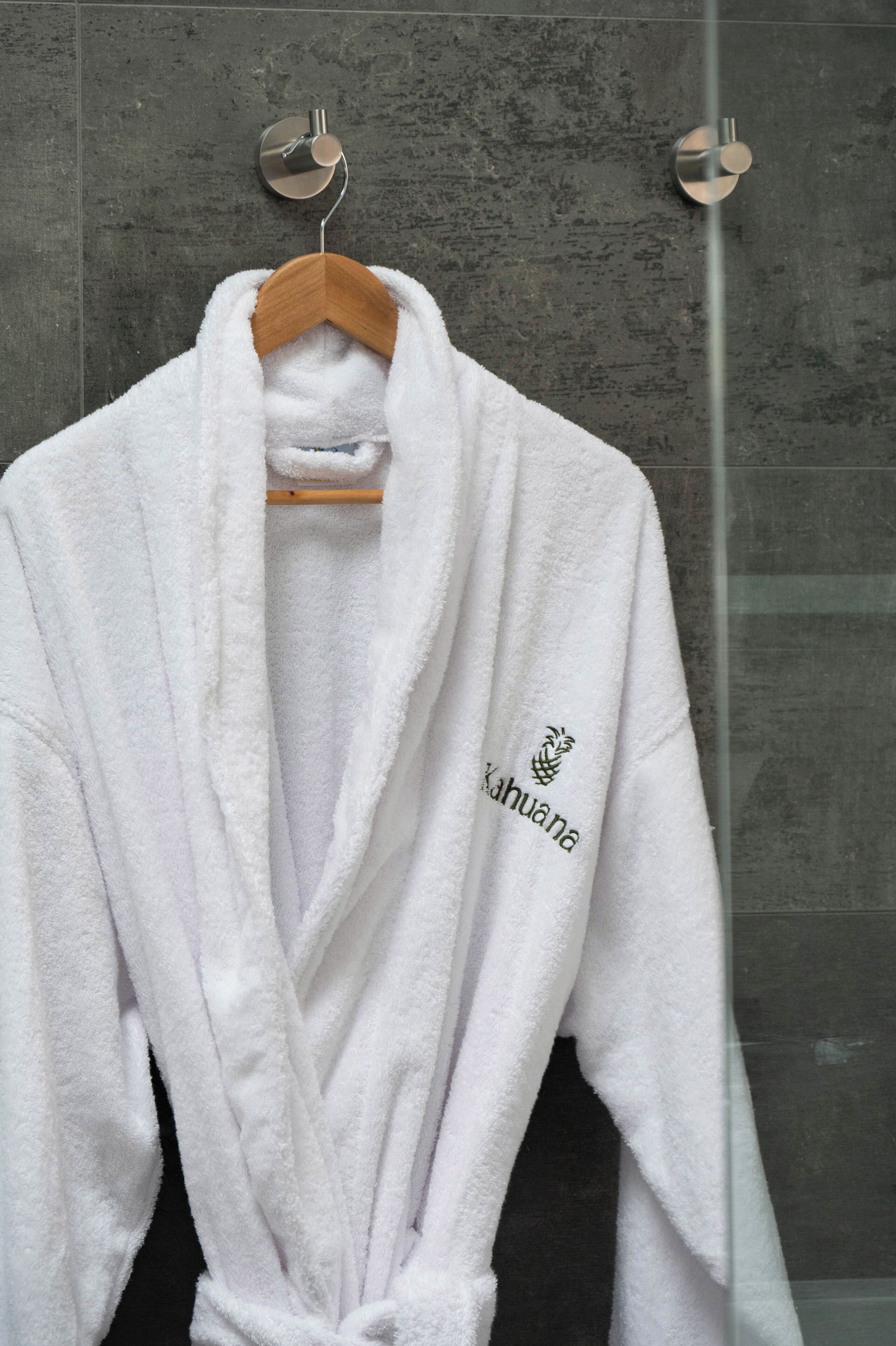 A white robe hanging on a wooden hanger in a bathroom with a textured wall and a glass shower door.