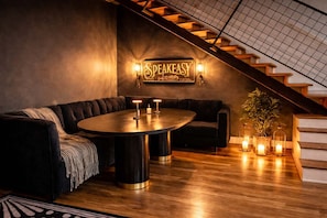 Un espacio lounge moderno con una mesa redonda, sofás de cuero oscuro, una escalera y un letrero que dice 'SPEAKEASY'.