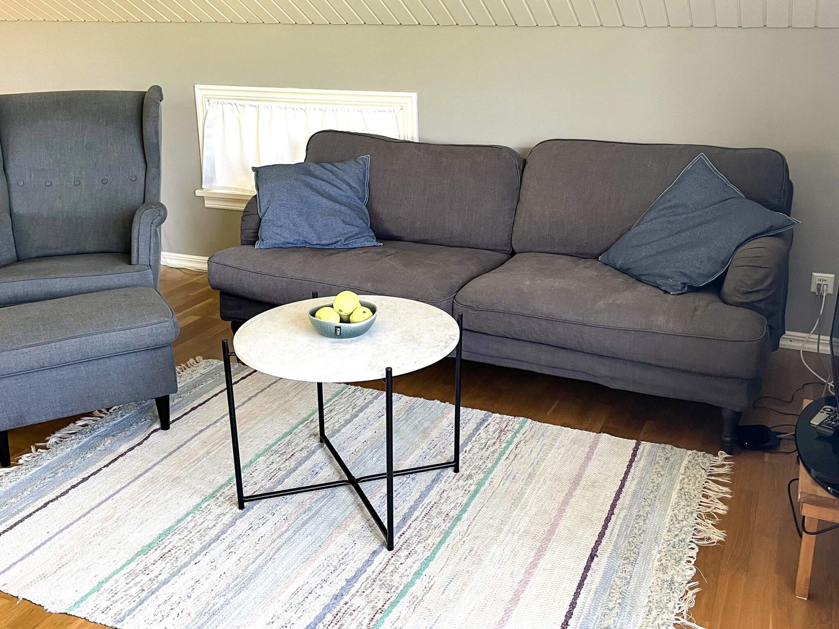 A living room with a grey sofa, a grey armchair, a round coffee table with a bowl of fruit, and a striped rug.