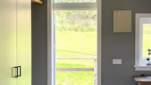Une pièce avec une porte donnant sur une terrasse en bois et une cour gazonnée. On y trouve une petite table avec une prise électrique et un interrupteur fixé au mur.