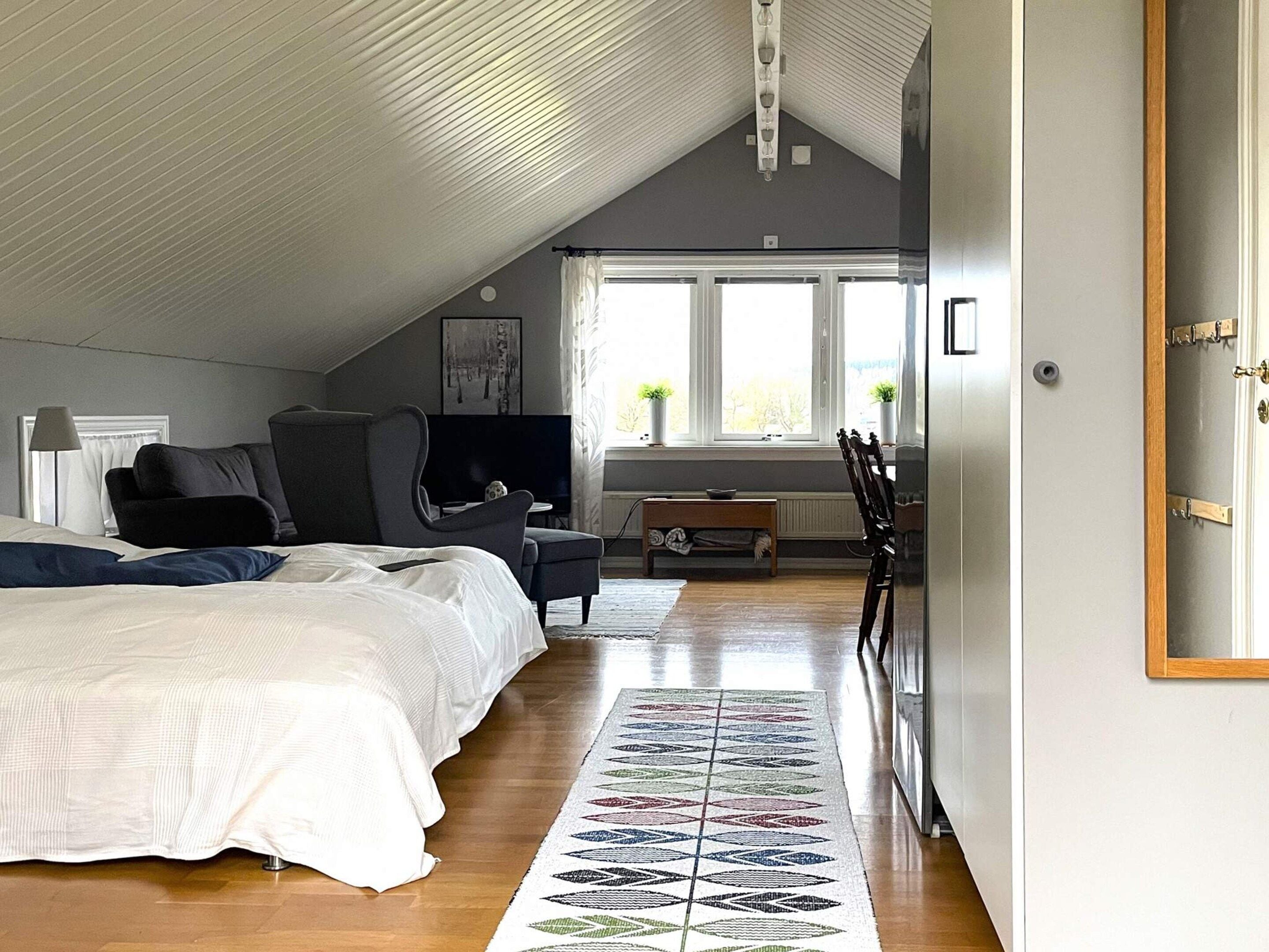 A bedroom with a sloped ceiling, a bed with white bedding, a grey armchair, a small table, and a window with curtains.