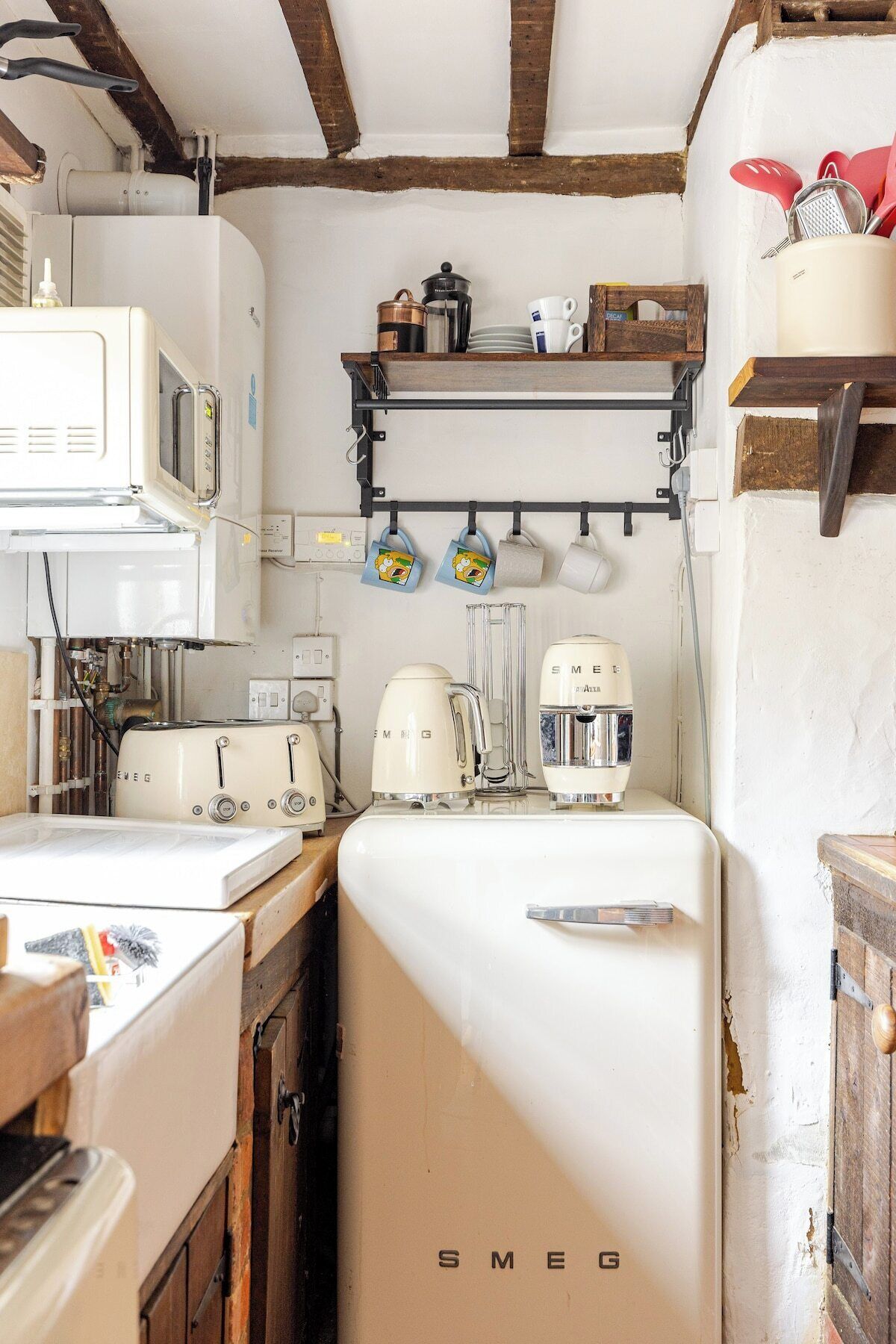 A kitchen with vintage appliances, including a white Smeg refrigerator, a toaster, and a coffee maker. There are hanging mugs and a shelf with kitchen utensils.