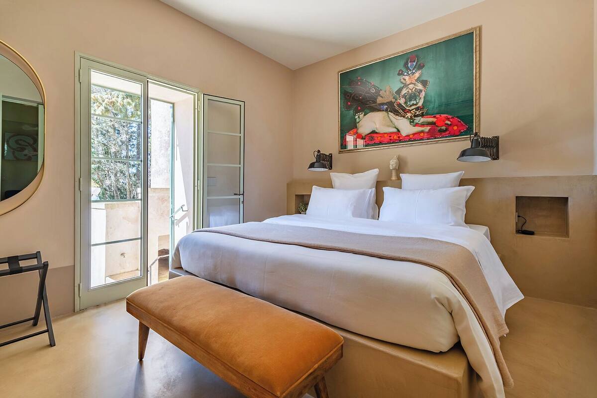 A bedroom with a large bed, a bench, a painting, and a view through a glass door.