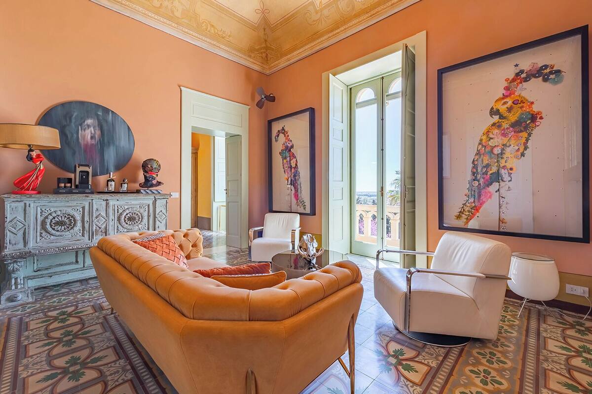 A spacious living room with a large sofa, a white armchair, and a patterned floor. There are decorative items on a cabinet and a painting on the wall.