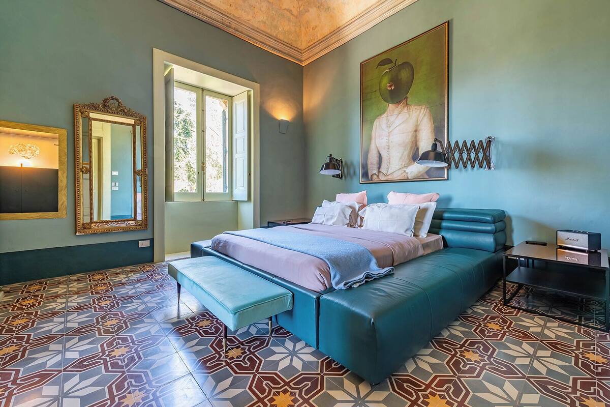 A bedroom with a large bed, a bench, a desk, and a painting on the wall.