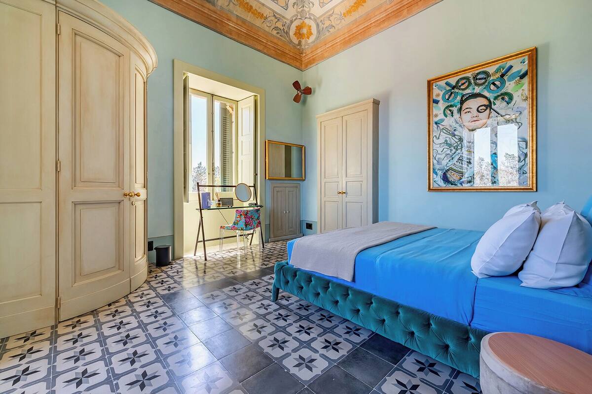 A bedroom with a blue bed, white pillows, a green upholstered bench, and a tiled floor with a geometric pattern.