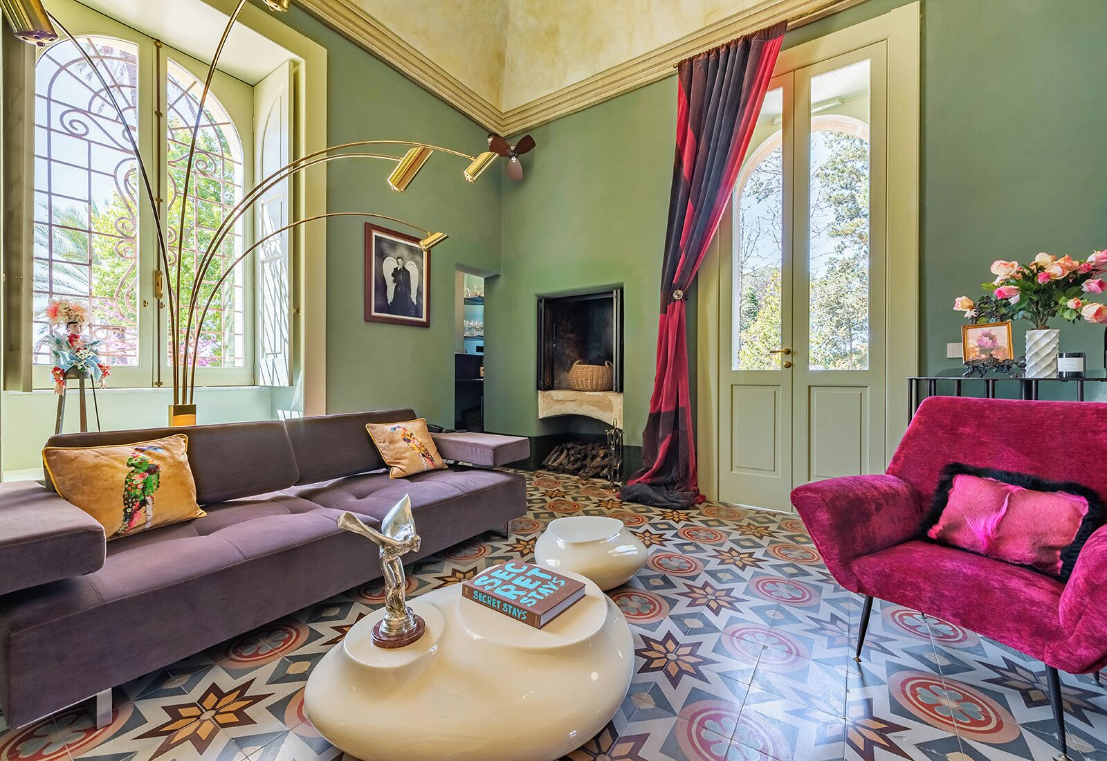 A room with a sofa, armchair, and a fireplace. There are decorative items on the fireplace mantel and a vase with flowers on a side table.
