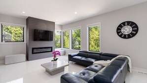 A modern living room with a black leather sofa, a flat-screen TV mounted on a wall, a fireplace, and large windows offering a view of greenery. - Brand-New 3Story Home with Rooftop in Midtown (Atlanta)