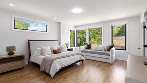 A modern bedroom with a large bed, a sofa, and a desk. There are multiple windows providing natural light and views of greenery. - Brand-New 3Story Home with Rooftop in Midtown (Atlanta)