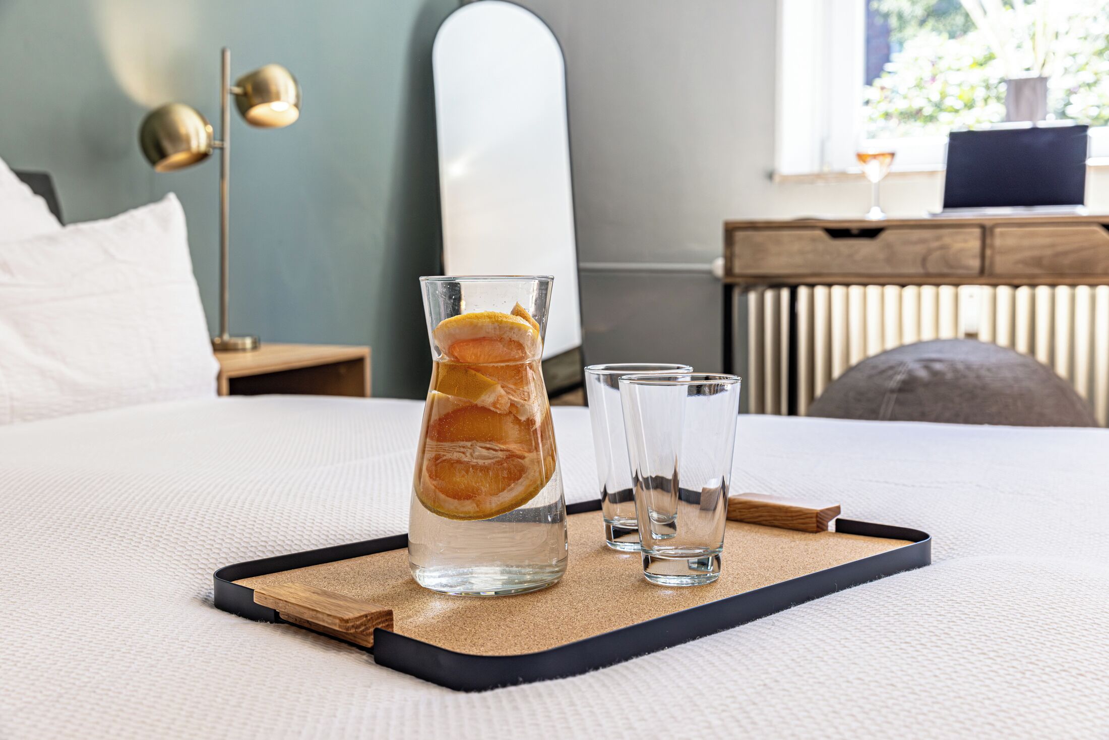 A bed with a tray holding a glass of water with lemon slices, two clear glasses, and a wooden tray.