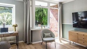 A modern living room with a wooden floor, a grey armchair, a wooden desk with a laptop, a radiator, a TV on a wooden stand, and a window with a view of greenery. - have-a-nice-stay - modern, near train station, quiet, 15 minutes by train to HH (Ahrensburg)