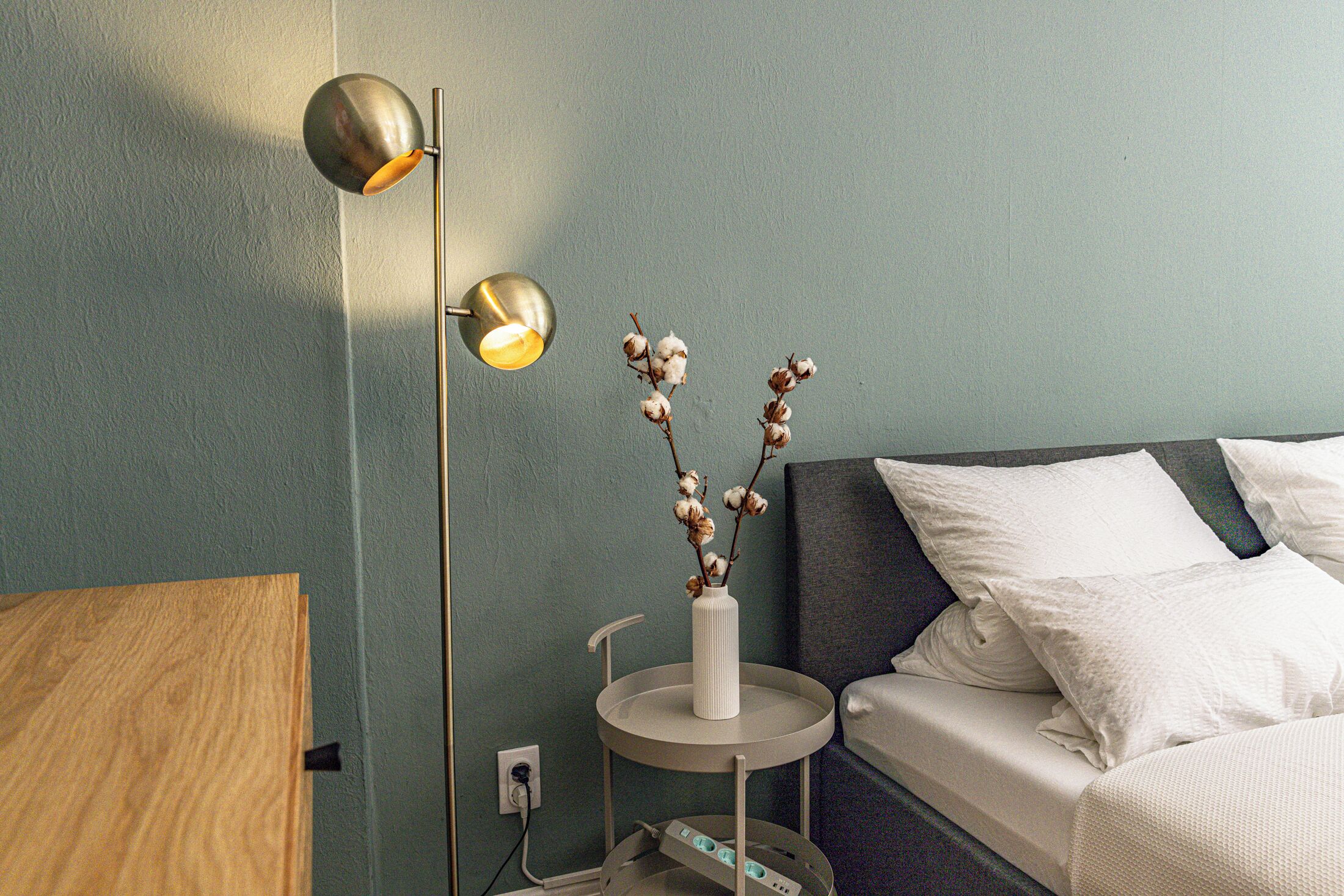Ett välordnat sovrum med en säng, ett sängbord, en lampa och en vas med torkade blommor.