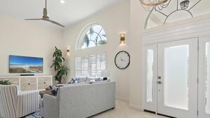 A spacious living room with a grey sofa, striped armchair, and a large TV. There is a ceiling fan, a plant, and a clock on the wall. - Waterfront 3BR retreat with heated pool, WiFi, and room for 8. (Marco Island)