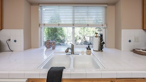 A kitchen with a double sink, white tiles, a window with blinds, and a view of greenery outside. - Eagle Retreat ~ Mountain Escape • New • With AC (Shaver Lake)