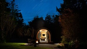 A cozy, circular, glass-enclosed structure with a wooden exterior, set in a garden at night. - Hideaway Cottages - Greenacres Estates (Tenby)