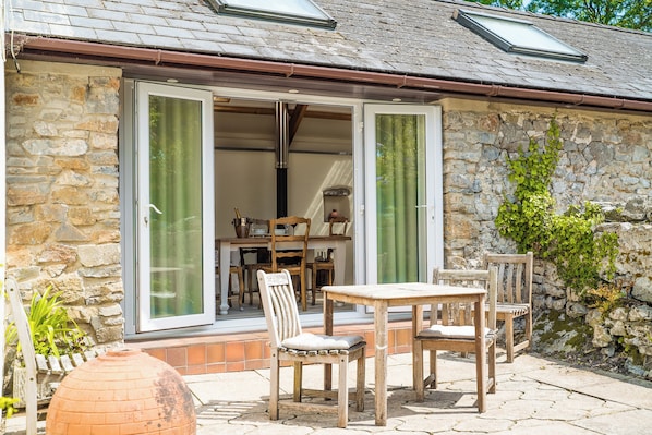 A stone building with a patio area featuring a wooden table and chairs, a brick oven, and a dining set. - Hideaway Cottages - Greenacres Estates (Tenby)