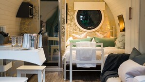 A cozy bedroom with a bed, a small table with glasses, and a window with a view of trees. - Hideaway Cottages - Greenacres Estates (Tenby)