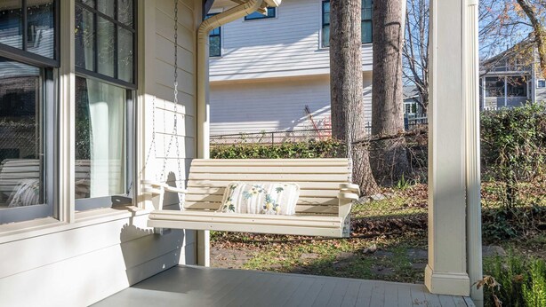 A porch with a wooden bench and a cushion, a white house in the background, and a tree to the right. - Ready Aim (Atlanta)