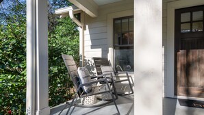 Une véranda meublée pour l’extérieur, comprenant deux fauteuils à bascule et une table, devant une maison dont la porte d’entrée et les fenêtres sont de couleur sombre.