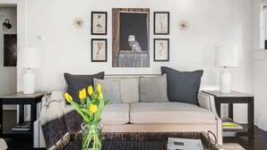 A living room with a sofa, two side tables, a vase with yellow flowers, framed botanical prints, and a painting of an owl. - Ready Aim (Atlanta)