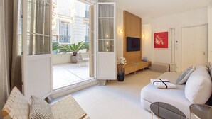 A modern living room with a sofa, a TV mounted on a wooden panel, a potted plant, and a view of the city through large windows. - N&J - PALMIERS TERRASSE - Central - Terrace (Niza)