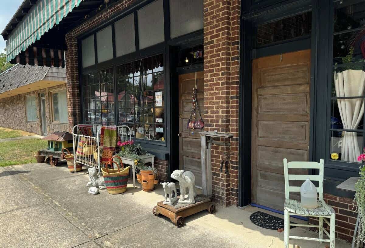 A storefront with large windows, a brick facade, and various items displayed outside, including a white chair and a statue.
