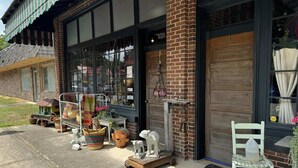 A storefront with large windows, a brick facade, and various items displayed outside, including a white chair and a statue.