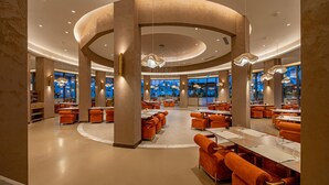 A spacious restaurant with round tables and orange upholstered chairs, featuring a central column and pendant lighting.