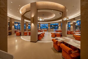 A spacious restaurant with round tables and orange upholstered chairs, featuring a central column and pendant lighting.