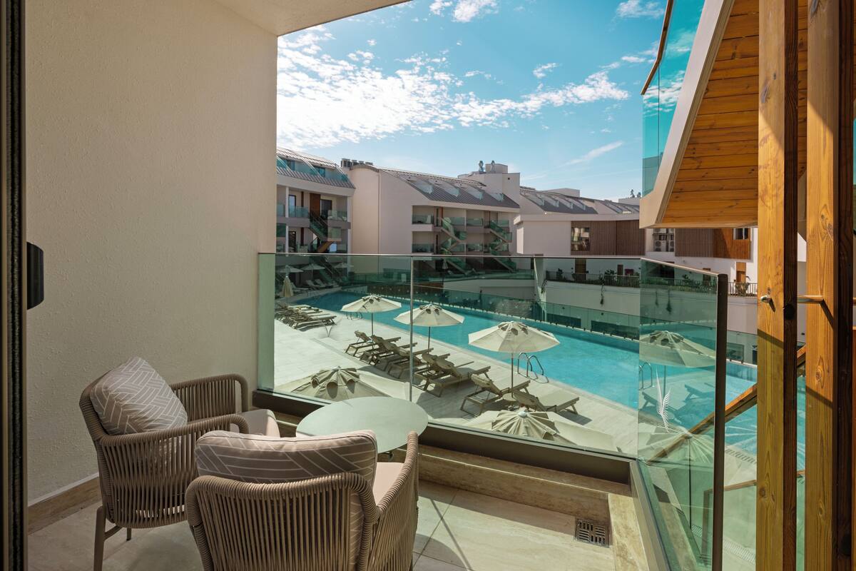 A balcony with wicker chairs and a glass table overlooking a pool area with umbrellas and lounge chairs.