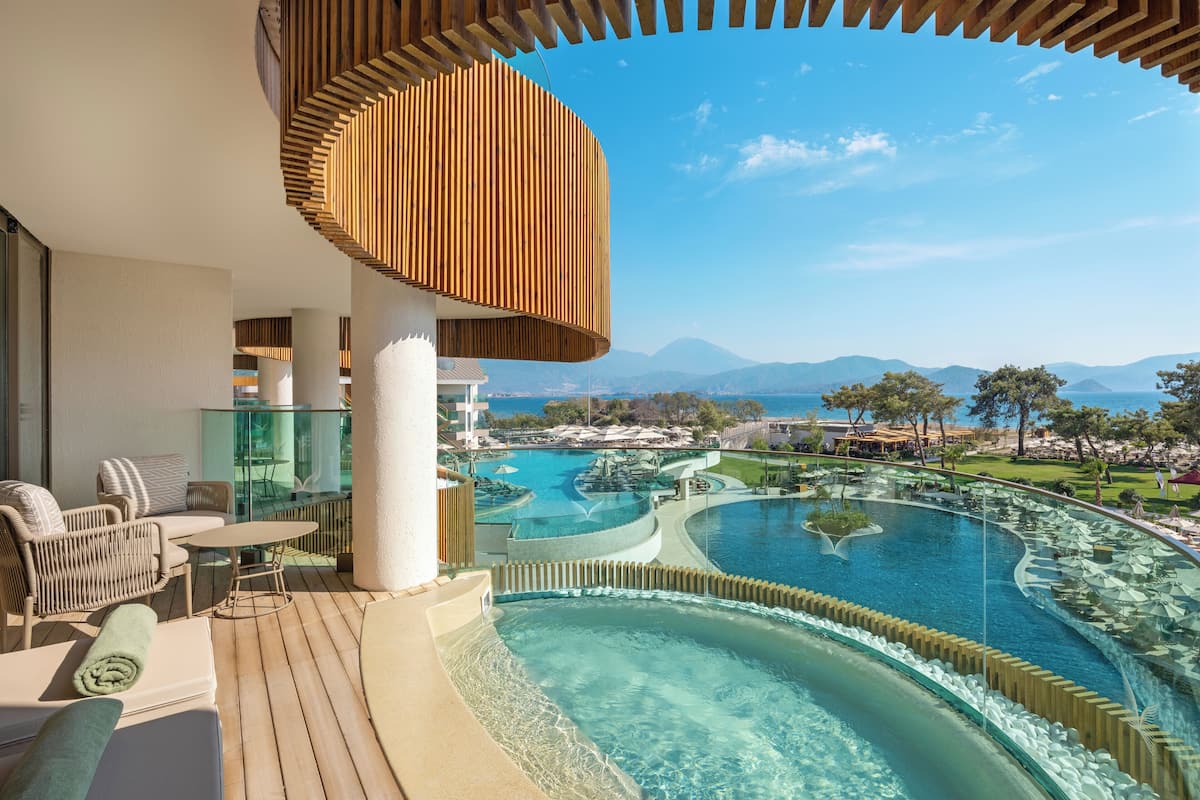 A balcony with wicker furniture, a glass table, and a view of a pool and mountains.