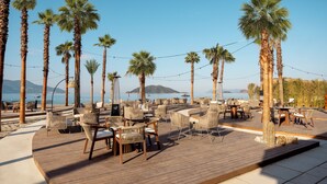Un espace extérieur aménagé avec des tables et des chaises en bois, situé au bord de la plage, entouré de palmiers et offrant une vue sur la mer.