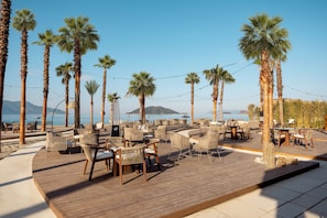 A beachside outdoor seating area with wooden tables and chairs, surrounded by palm trees, overlooking the sea. - Liberty Signa - All inclusive (Fethiye)