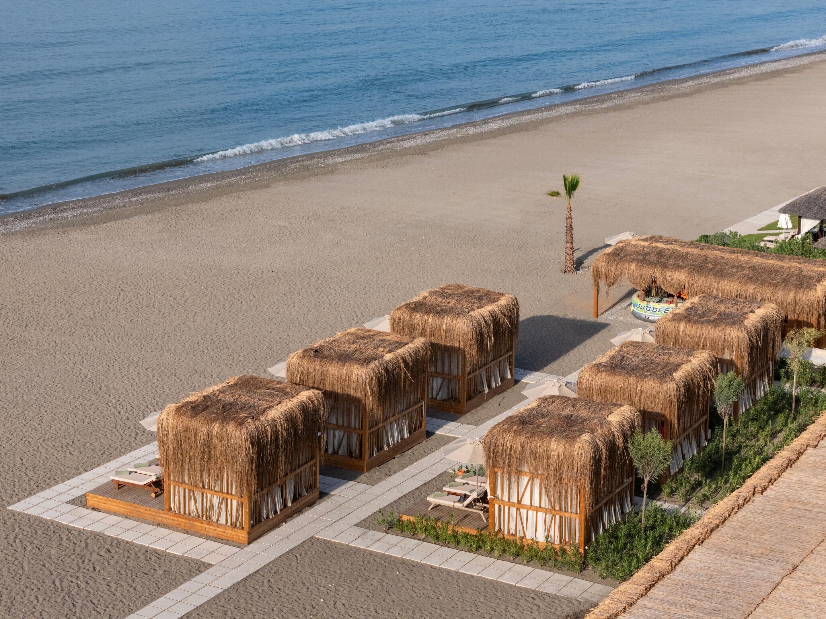A beachside resort with thatched-roof huts arranged in rows, each with a small garden area and a view of the sea.