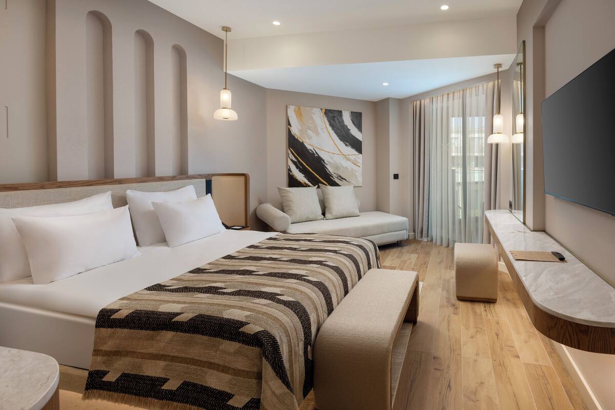 A modern hotel room with a large bed, a sofa, a bench, and a desk with a flat-screen TV.