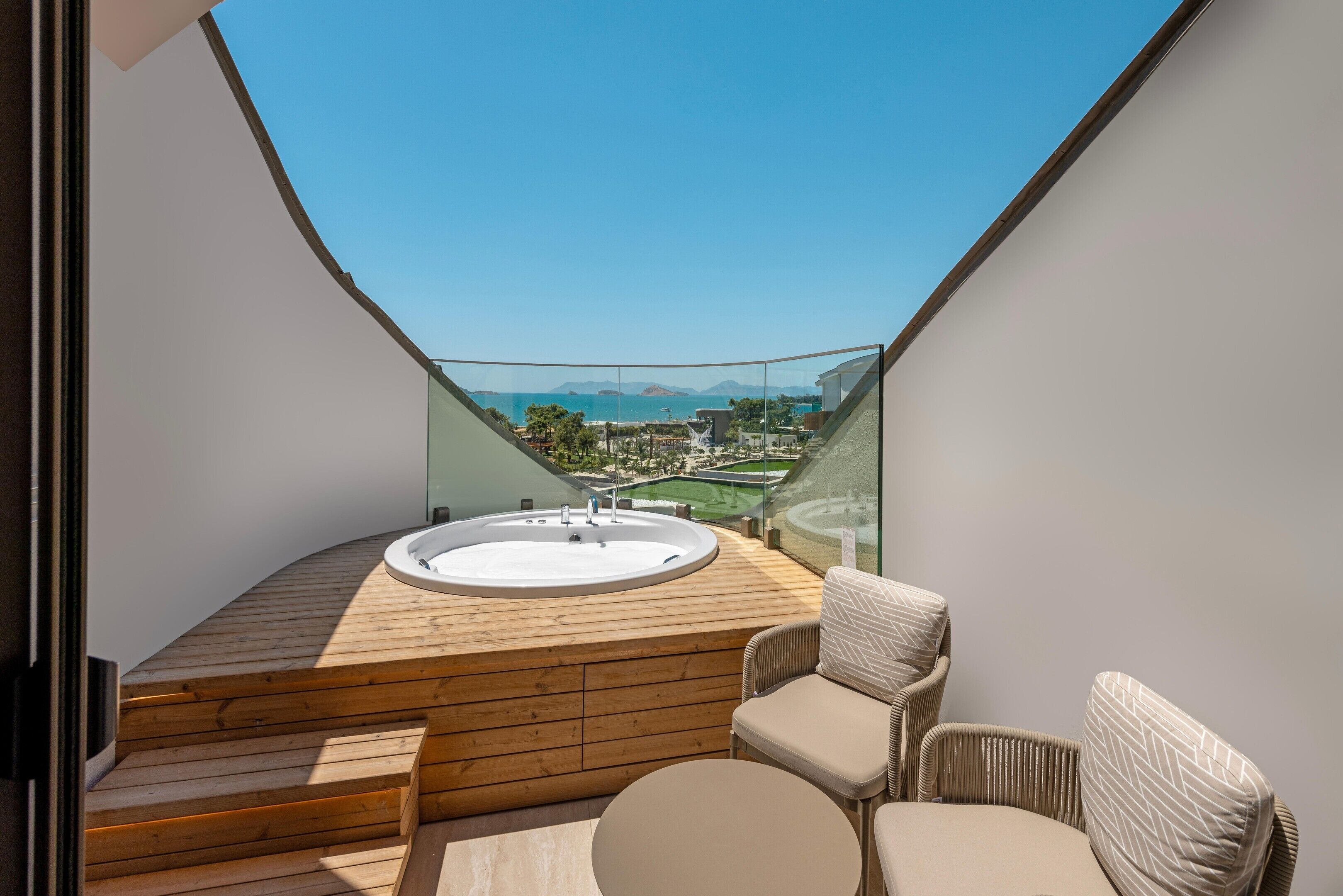 A balcony with a hot tub, wooden deck, and seating area with a view of the sea and mountains.