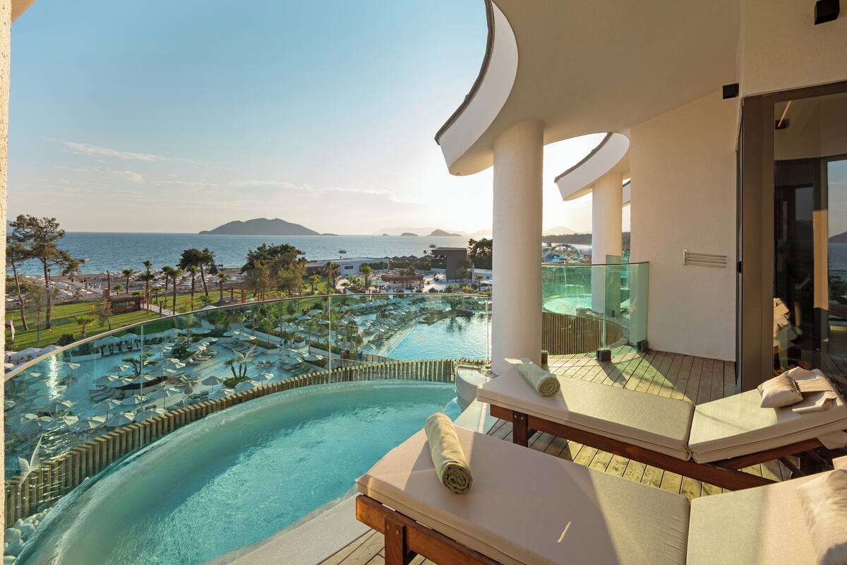 A poolside lounge with sun loungers, a view of a resort pool, and a distant beach and mountains.