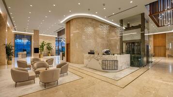 A modern hotel lobby with a reception desk, seating area, and a glass elevator.