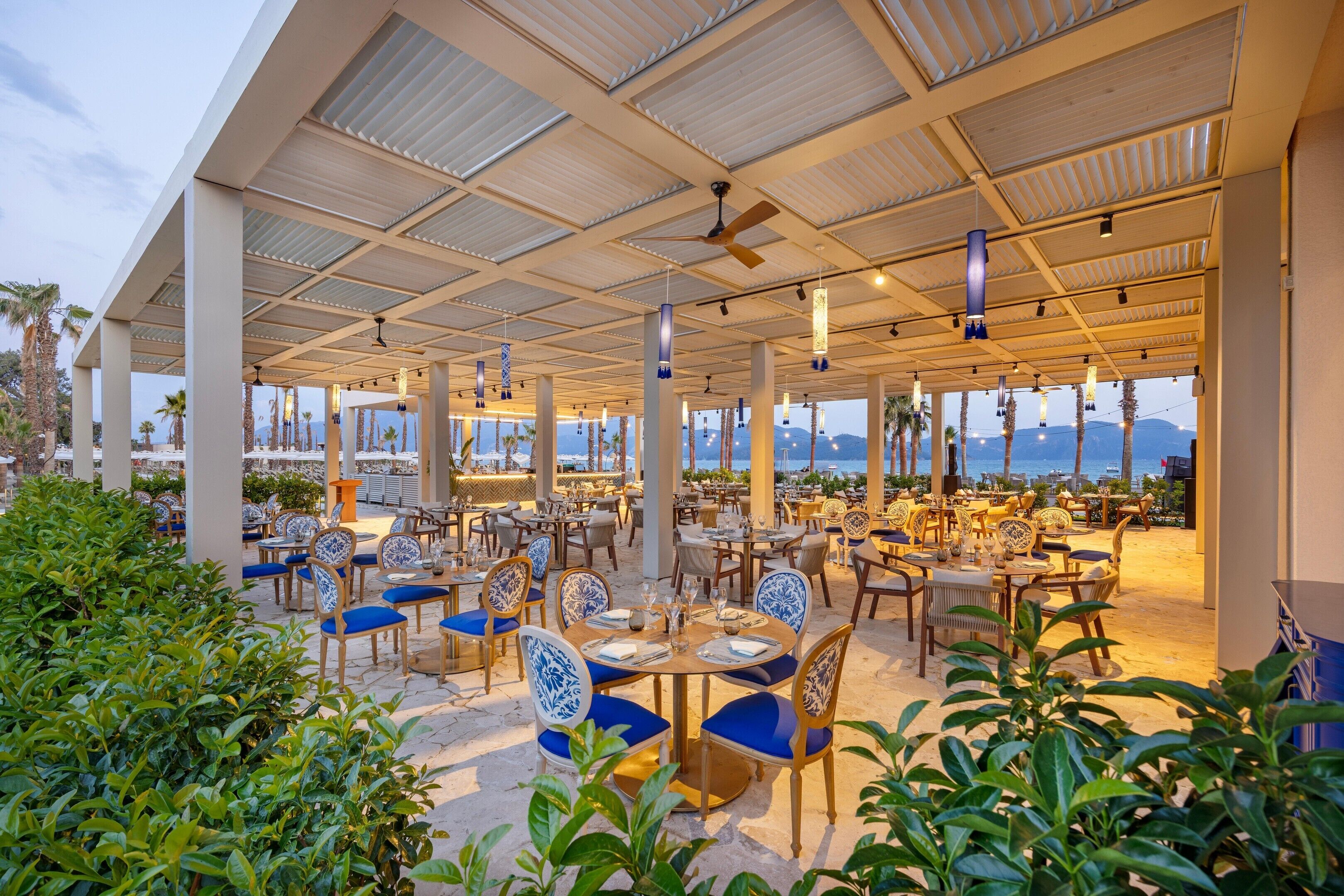 An outdoor dining area with tables and chairs, a covered roof, and a view of the sea.