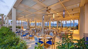 An outdoor dining area with tables and chairs, a covered roof, and a view of the sea.