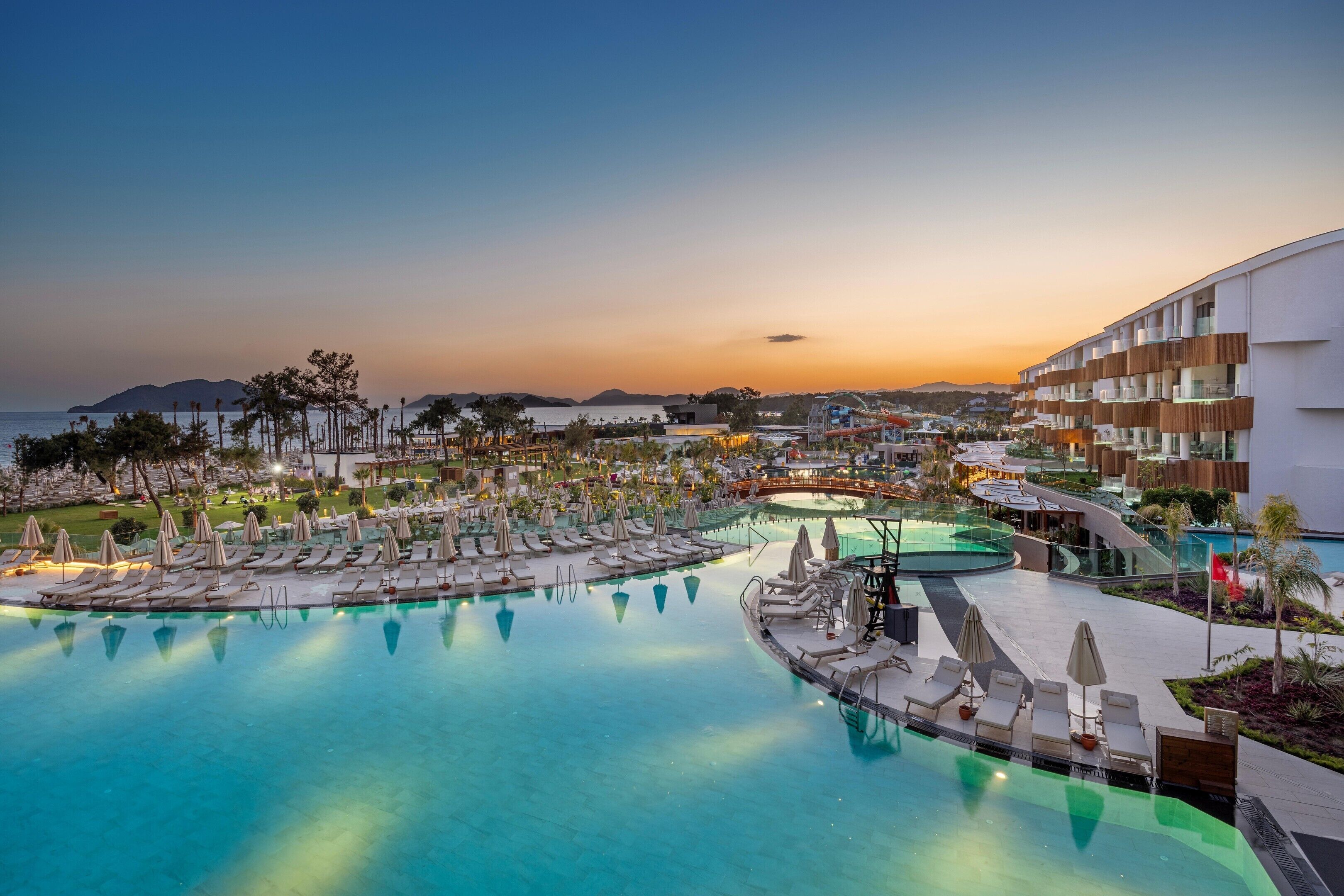 A resort with a large pool, lounge areas, and a view of the sea at sunset.